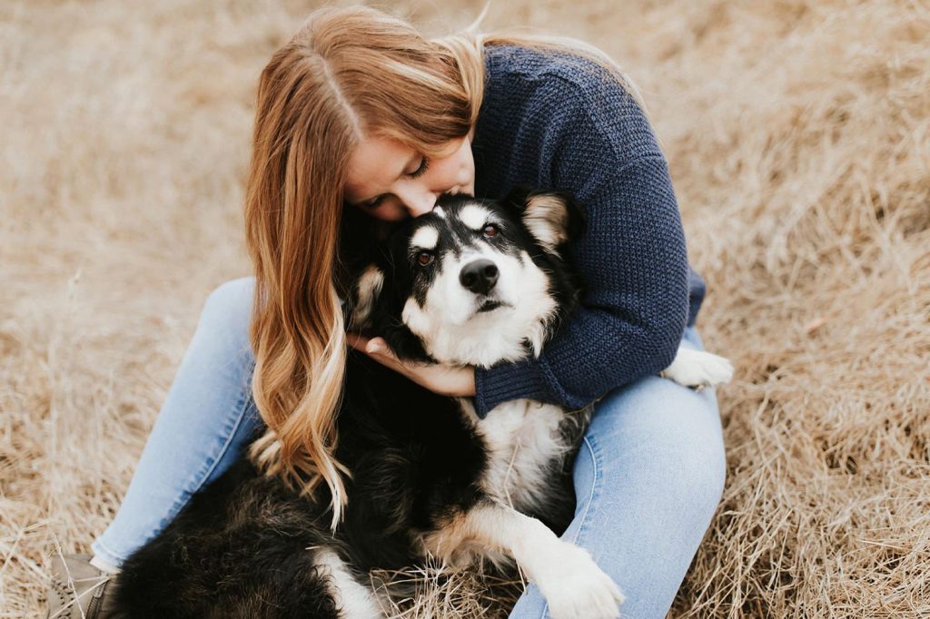 A person with long brown hair hugs a black and white dog while sitting on dry grass in an open field.