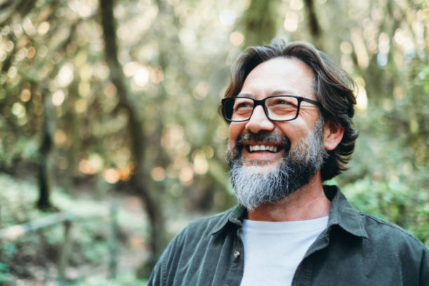 A man with glasses and a gray beard smiles broadly while standing in a lush forest with trees and greenery in the background.